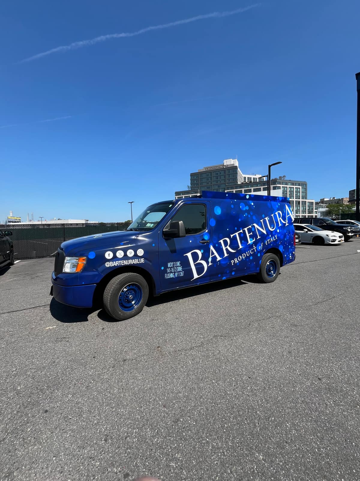 Side view of the blue Nissan NV cargo van wrapped for Bartenura, parked in a retail plaza parking lot