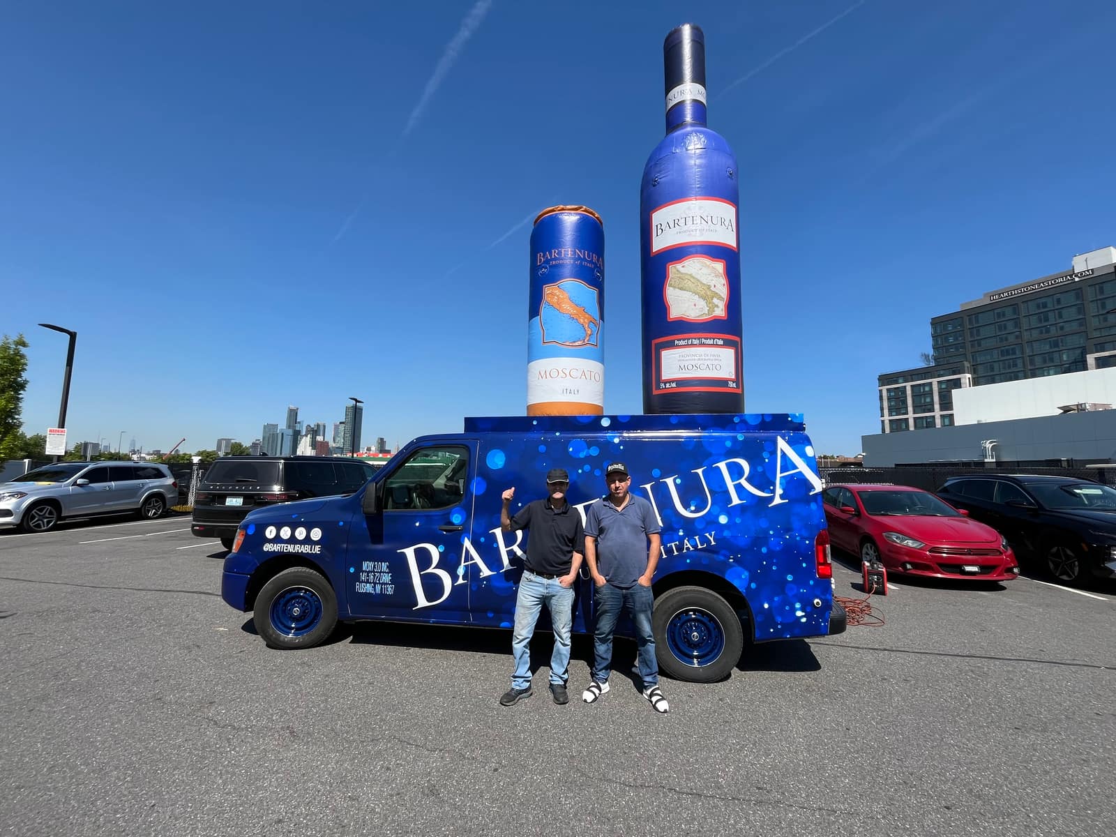 Blue Bartenura-wrapped Nissan NV van with two people standing beside it and a giant inflatable wine bottle behind, in a retail plaza parking lot