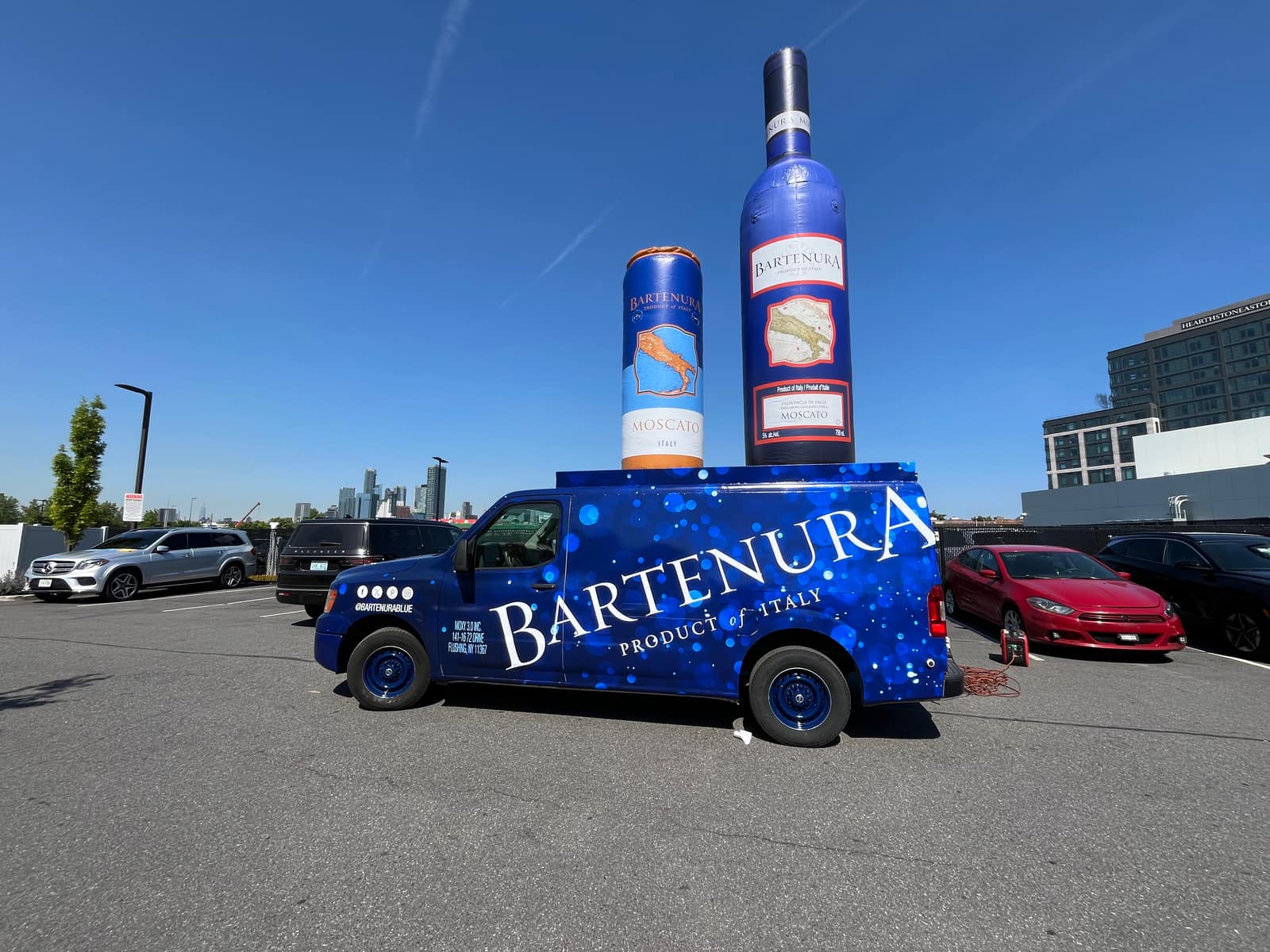 Blue Bartenura-wrapped Nissan NV van parked beside a giant inflatable wine bottle in a retail plaza parking lot