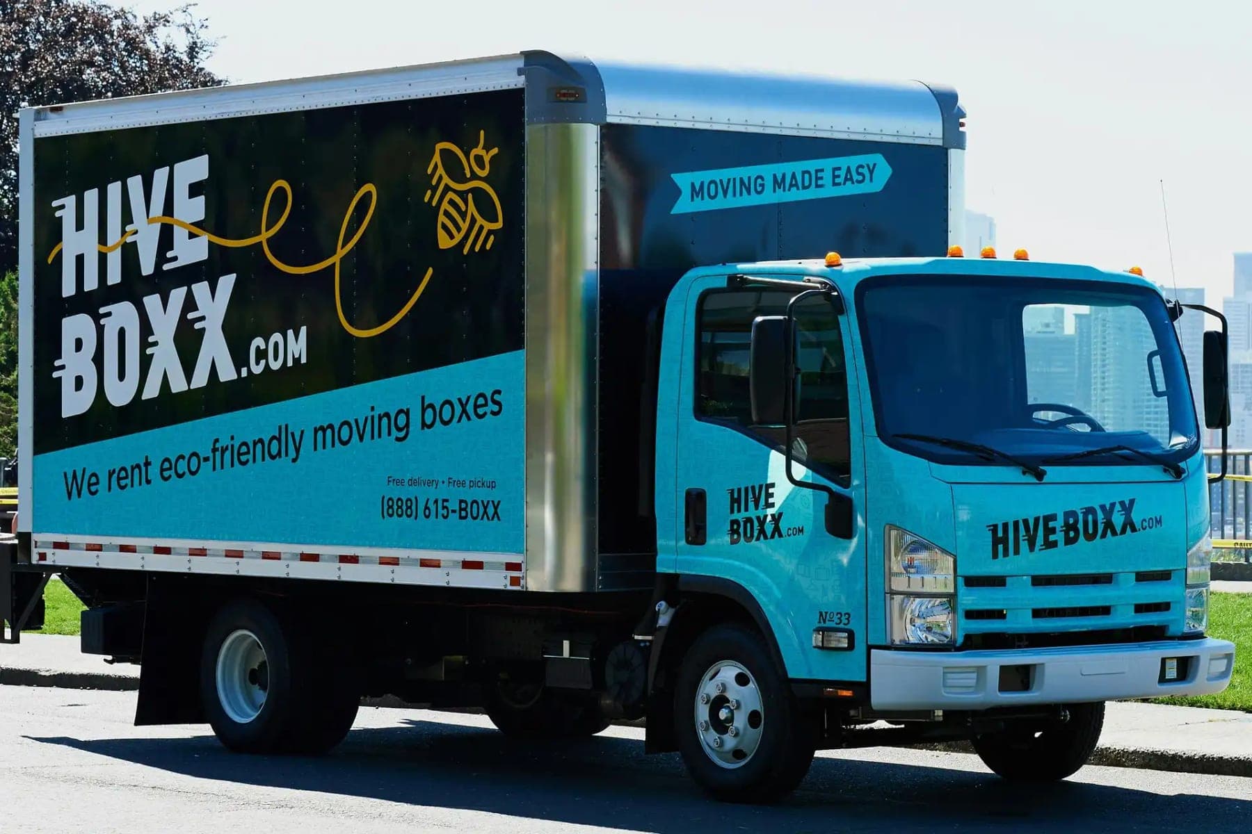 Teal Isuzu box truck for HiveBoxx.com with a dark box graphic, bee illustration and We rent eco-friendly moving boxes lettering, side view on a street