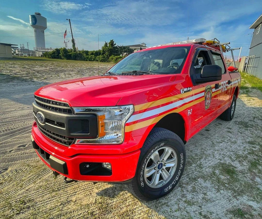 Red Ford F-150 crew cab with white and gold reflective stripe graphics, FIRE lettering and a fire-department seal on the door, front three-quarter view on sand