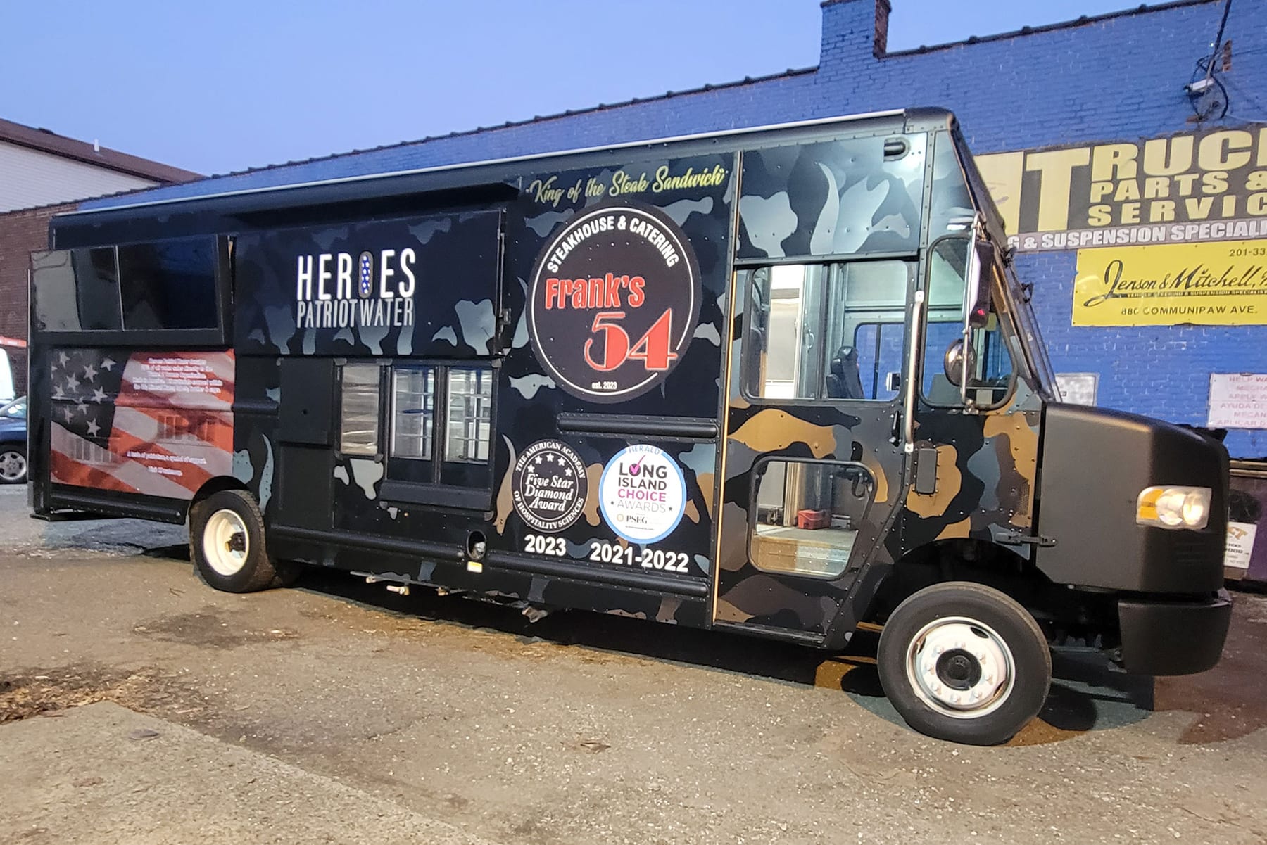 Black Heroes cheesesteak truck with camo accents, parked at a truck service yard